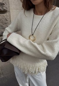 Belo, teksturirano pulover s raztrganim robom, nosi se z črnim ogrlico in rjavim nahrbtnikom. Nahrbtnik ima strukturno zasnovo in gladko usnjeno površino.