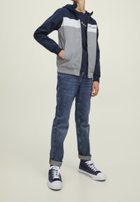 Grå och marinblå färgblockerad zip-up jacka med huva, kombinerad med blå denimjeans och marinblå sneakers, med vita snören och en slät yta.