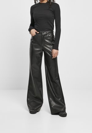 Kobieta ubrana w czarną bluzkę z długim rękawem z falbankami na końcach i wysokie, szerokie spodnie ze skóry, stojąca z jedną ręką w kieszeni.