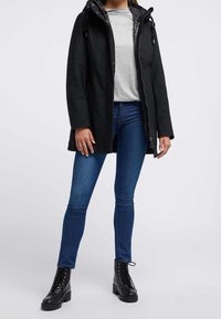 Chaqueta negra impermeable con cierre de cremallera, capucha y cordones ajustables, llevada sobre una camiseta gris y jeans azules, combinada con botas de combate negras.