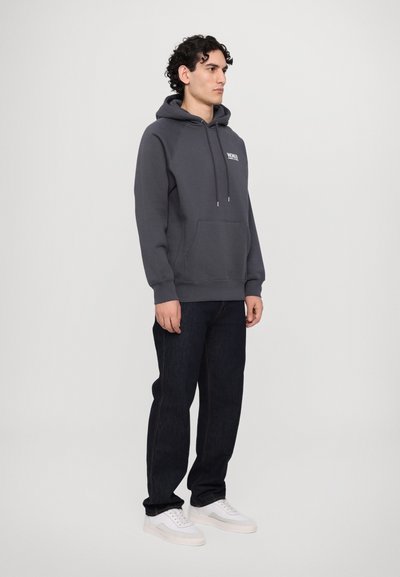 Donkergrijze hoodie met een voorzak, een capuchon met trekkoord en een wit logo. Gecombineerd met donkerblauwe jeans en witte sneakers.