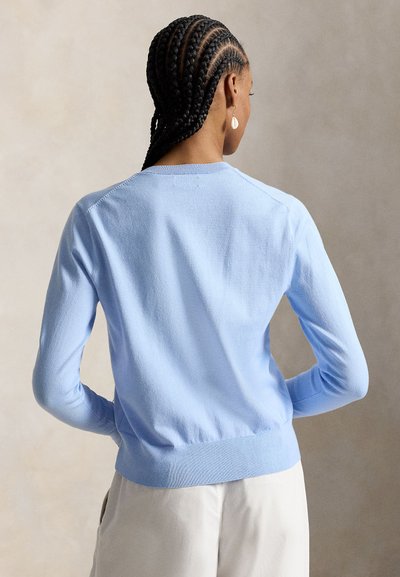 Frau mit geflochtenem Haar, die einen hellblauen Pullover mit langen Ärmeln und eine weiße Hose trägt, von hinten vor einem schlichten Hintergrund gezeigt.