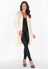 Egy nő krémszínű hosszú kardigánt visel, amelyen flitteres részletek találhatók, fekete V-nyakú felsőt, fekete magas derekú leggingset és fekete magassarkú szandálokat, miközben pózol.