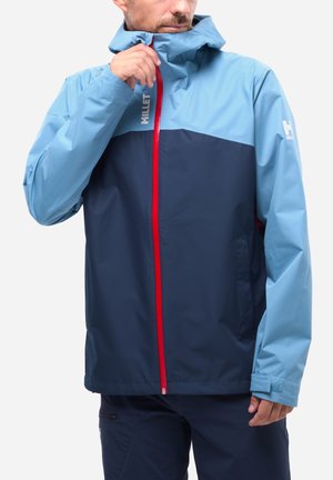 Mann trägt zweifarbig blaue wasserdichte Jacke mit rotem Reißverschluss, richtet den Kragen, steht vor schlichtem Hintergrund.