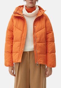 Vrouw in een feloranje pufferjack over een crèmekleurige coltrui en tan wijde broek tegen een effen achtergrond.