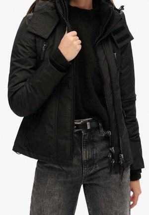 Woman wearing a black puffer jacket with multiple zippers, black sweater, and faded black jeans, holding the jacket's collar with one hand.