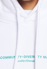 Felpa bianca con cappuccio caratterizzata da testo teal sul davanti: "LA COMUNITÀ E LA DIVERSITÀ IMPORTANO" e "uguaglianza= cambiamento". Tessuto morbido con cappuccio regolabile.