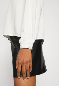 Main droite d'une personne portant une blouse blanche avec un poignet boutonné et une jupe en cuir noir sur un fond uni.