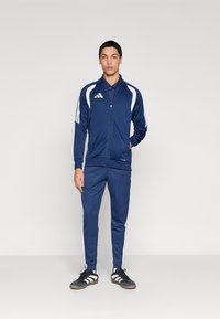 Σκουρόχρωμο μπλε σετ φόρμας από απαλό, ελαφρύ ύφασμα, με λευκές ρίγες στα μανίκια και στα πόδια του παντελονιού, και μπουφάν με φερμουάρ.