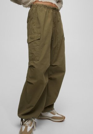 Luźne oliwkowozielone spodnie typu cargo z dużymi bocznymi kieszeniami, ściągaczem w talii i elastycznymi mankietami, noszone z białymi i beżowymi sneakersami.