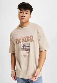 Beige bomulds T-shirt med korte ærmer, med et grafisk print af et palmetræ og teksten "AUTORISERET FORHANDLER". Casual fit.