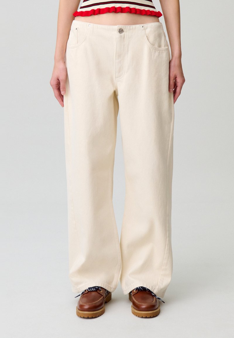 Pantalones de pierna ancha color crema hechos de algodón con una textura suave, que incluyen bolsillos frontales y cierre de botón, combinados con zapatos marrones.
