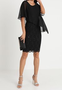 Svart klänning med dubbla lager som har ett genomskinligt, flödande överdrag och en paljetterad pennkjol. Matchad med silvriga remsandaler och en svart kuvertväska.