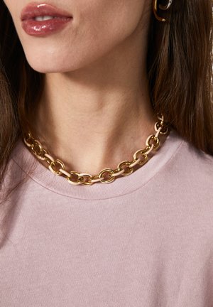 Goldene Kettenhalskette mit ovalen Gliedern, getragen über einem hellrosa gerippten Shirt. Glatte Textur und ein einfaches, markantes Design.