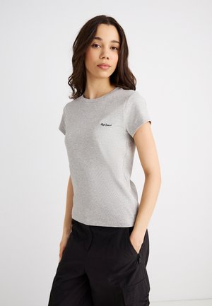 Fiatal nő hosszú sötét hajjal, világosszürke rövid ujjú, kerek nyakú pólót és fekete cargo nadrágot viselve, zsebeibe tett kézzel áll.