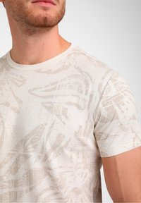 T-shirt beige à manches courtes en tissu doux, présentant un motif abstrait texturé et une petite étiquette logo sur la poitrine.