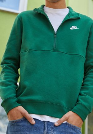 Osoba ubrana w zieloną bluzę Nike z półzamkiem na białej koszuli, ręce częściowo w kieszeniach, stojąca na tle rozmytego zielonego tła.