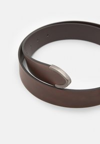 Cintura di pelle marrone arrotolata con fibbia a forma di ferro di cavallo incisa color argento su sfondo bianco.