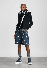 Pull en tricot noir avec des boutons, short bleu marine avec broderie florale blanche, chaussettes grises et baskets montantes bleues avec semelles blanches.
