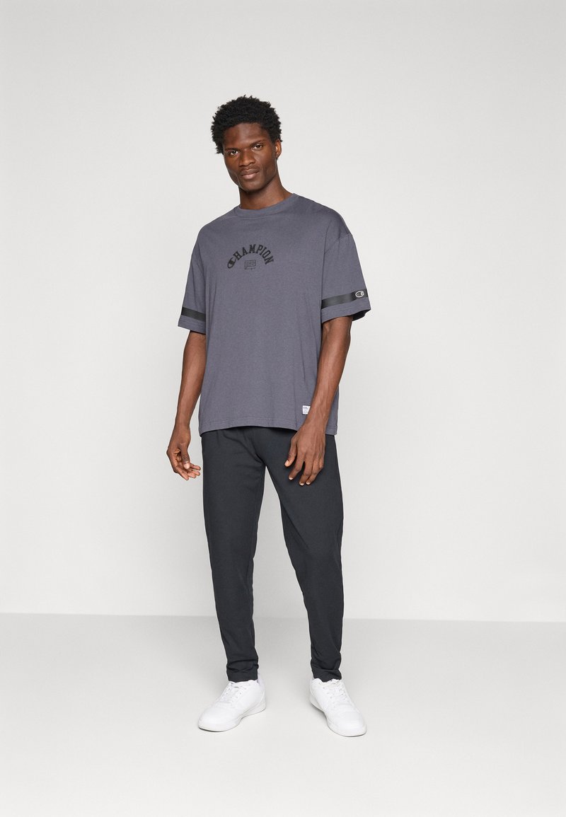 Graues T-Shirt mit kurzen Ärmeln und schwarzem Logo, ausgestattet mit Satinakzenten an den Ärmeln. Kombiniert mit dunklen Jogginghosen und weißen Sneakers.
