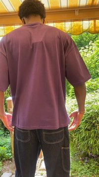 Vijolična bombažna oversized majica s krojenim ovratnikom, vidna s hrbtne strani. V kombinaciji s temnimi jeans hlačami, ki imajo zadnje žepe in kontrastno šiv.