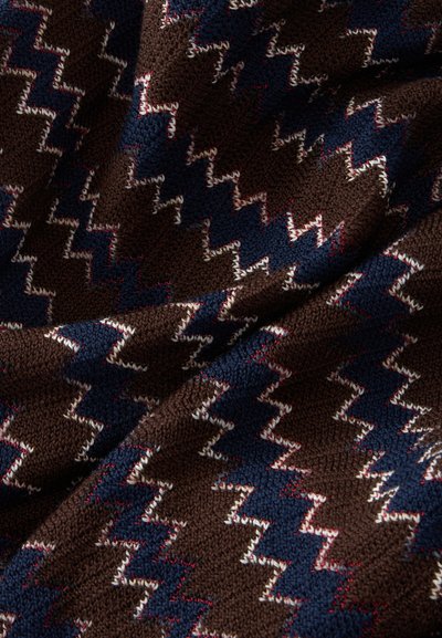 Gros plan d’un tissu tricoté marron avec un motif en zigzag bleu marine, blanc et des accents rouges, montrant la texture et les plis.