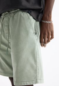 Shorts vert clair avec une taille élastique, un cordon de serrage ajustable, des poches latérales et une texture lisse et mate. Étiquette logo noire visible.