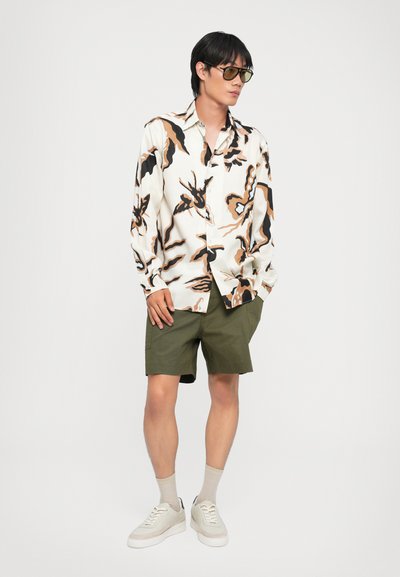 Langärmliges Hemd mit weißem Grundton, versehen mit abstrakten Mustern in Schwarz und Braun, kombiniert mit olivgrünen Shorts und weißen Sneakers.