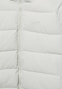 Doudoune grise claire avec fermeture éclair verticale, motif matelassé horizontal et logo brodé sur la poitrine. Texture lisse.