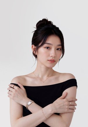 Nuori nainen, jolla on hiukset nutturalla, pukeutunut olkapäät paljastavaan mustaan toppiin, kultakelloon, sormuksiin, hennon kaulakoruun ja rannekoruun, neutraalia taustaa vasten.