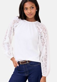 Valge pluus pitsiste varrukatega, tekstuurse kangaga, ümmarguse kaelusega ja kogutud õlalehvikuga, kombineeritud tumesiniste teksade ja pruuni vöökohaga.