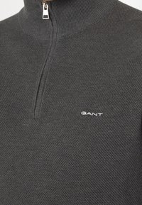 Pull en maille texturée gris foncé avec une fermeture éclair argentée à mi-hauteur d'un col montant et un logo blanc "GANT" sur la poitrine.