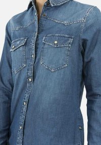 Camicia di denim di tonalità blu medio, con colletto a punta, chiusura frontale con bottoni e due tasche sul petto con pattine. Texture liscia.