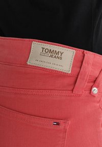 Jeans de mezclilla rojos con una etiqueta de cuero marcada "TOMMY JEANS" y "UN ORIGINAL AMERICANO" cerca de la cintura, con un botón de metal y bolsillos.