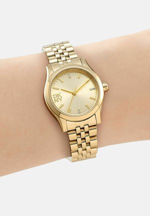 Goudkleurig DKNY-horloge met schakelen armband, ronde wijzerplaat, minimale uurmarkeringen en drie wijzers, gedragen om de pols van een persoon.