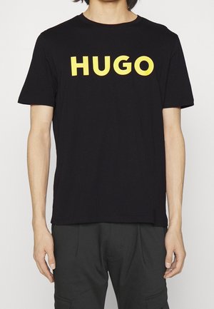 Zwart T-shirt met korte mouwen en een opvallende gele "HUGO" tekst op de borst, gedragen door een persoon in een donkere broek tegen een effen witte achtergrond.
