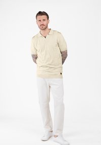 Uomo con tatuaggi che indossa una polo a maniche corte lavorata beige, pantaloni bianchi e sneakers bianche, in piedi con le mani dietro la schiena su sfondo bianco.