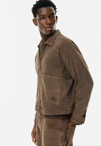 Brun denimjakke med lynlås foran, spidse krave og to forlommer. Har en ribbet kant og manchetter med en falmet tekstur.