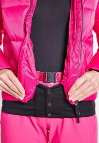 Veste isolante rose vif avec fermeture zippée, associée à une ceinture noire ajustable. Dotée de boutons-pression et d'un tissu contrastant.