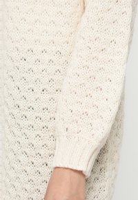 Gros plan d'une main portant une manche de pull en tricot de couleur crème avec un poignet côtelé et un motif texturé.