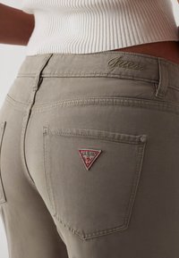 Blugi din denim khaki, cu un design ajustat, prezentând un mic patch triunghiular roșu și alb pe buzunarul din spate și detalii delicate de cusătură.