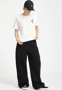 Hvid bomulds t-shirt med et navyblåt grafisk design på venstre bryst, kombineret med løstsiddende sorte bukser og en sort kasket.