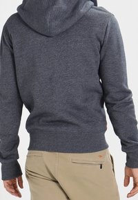 Hombre vistiendo un sudadera con capucha gris oscuro y pantalones beige, visto desde atrás con los brazos relajados a los lados.