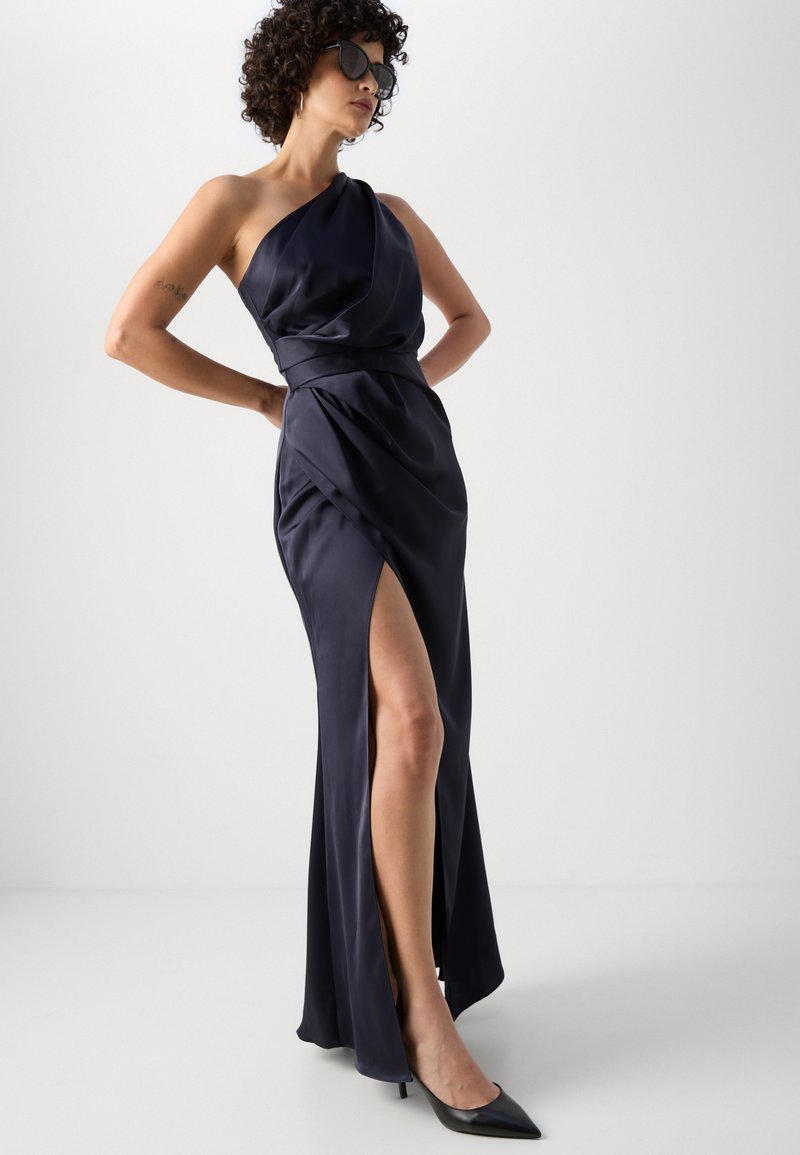 Granatowa satynowa sukienka z jednym ramiączkiem i bocznym rozcięciem, z drapowanym materiałem i kontrastującym paskiem w talii. Połączona z czarnymi szpilkami.