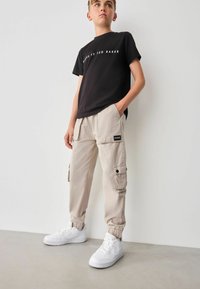 Schwarzes Baumwoll-T-Shirt mit weißem Text, beige Cargo-Hosen mit Seitentaschen und schwarzem Markenpatch, kombiniert mit weißen Sneakers.