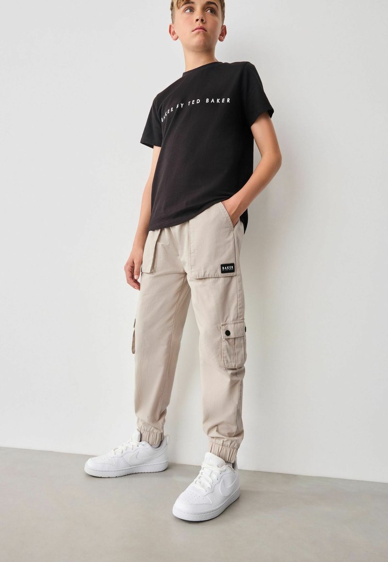 Schwarzes Baumwoll-T-Shirt mit weißem Text, beige Cargo-Hosen mit Seitentaschen und schwarzem Markenpatch, kombiniert mit weißen Sneakers.