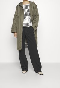 Parka verde oliva con cappuccio, chiusura con zip e tasche laterali. Sotto, una maglietta a righe bianche e blu navy. Pantaloni neri larghi e scarpe beige.