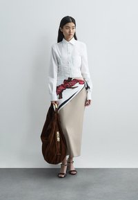 Bijela košulja s gumbima i detaljima od naborane tkanine, bež suknja sa šarenim šalom, smeđa suknja torba od antilopa i crne sandale s remenčićem oko gležnja.