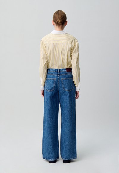 Camisa a rayas amarillas con cuello blanco, combinada con jeans de mezclilla azul de pierna ancha que cuentan con bolsillos traseros y una etiqueta de cuero.