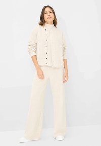 Beige strikket jakke med trykknapper, oversized pasform, ledsaget af matchende brede bukser i et blødt, struktureret stof. Hvide sneakers.